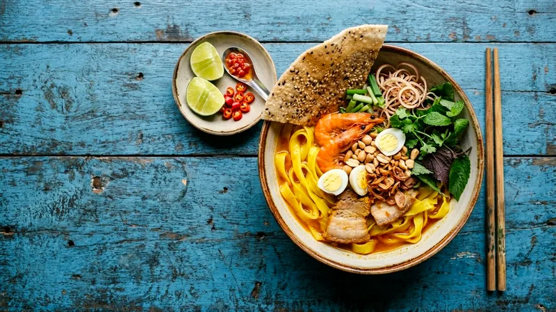 Bowl of mi quang noodles with shrimp, herbs and rice cracker on blue table